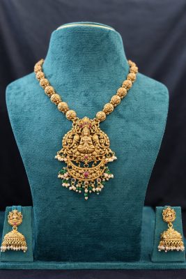 Embrace divine elegance with this stunning Lakshmi Temple Jewelry Set, a timeless piece that reflects the rich heritage of South Indian craftsmanship. The necklace features a chain of intricately carved Nakshi gold beads in a beautiful matte antique finish, leading to a magnificent Goddess Lakshmi pendant. The pendant showcases detailed temple artistry with the deity seated gracefully, surrounded by elaborate floral scrollwork. Ruby-colored stone accents add a touch of color, while delicate pearl clusters and green bead drops create a charming finishing touch. The matching jhumka earrings feature the same Lakshmi motif with traditional bell-shaped drops adorned with pearls, completing this divine ensemble.