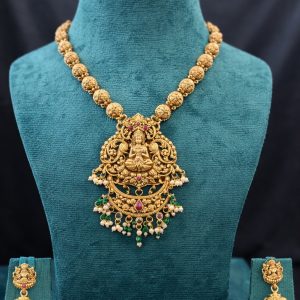 Embrace divine elegance with this stunning Lakshmi Temple Jewelry Set, a timeless piece that reflects the rich heritage of South Indian craftsmanship. The necklace features a chain of intricately carved Nakshi gold beads in a beautiful matte antique finish, leading to a magnificent Goddess Lakshmi pendant. The pendant showcases detailed temple artistry with the deity seated gracefully, surrounded by elaborate floral scrollwork. Ruby-colored stone accents add a touch of color, while delicate pearl clusters and green bead drops create a charming finishing touch. The matching jhumka earrings feature the same Lakshmi motif with traditional bell-shaped drops adorned with pearls, completing this divine ensemble.