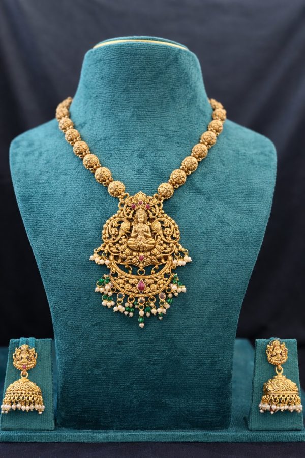 Embrace divine elegance with this stunning Lakshmi Temple Jewelry Set, a timeless piece that reflects the rich heritage of South Indian craftsmanship. The necklace features a chain of intricately carved Nakshi gold beads in a beautiful matte antique finish, leading to a magnificent Goddess Lakshmi pendant. The pendant showcases detailed temple artistry with the deity seated gracefully, surrounded by elaborate floral scrollwork. Ruby-colored stone accents add a touch of color, while delicate pearl clusters and green bead drops create a charming finishing touch. The matching jhumka earrings feature the same Lakshmi motif with traditional bell-shaped drops adorned with pearls, completing this divine ensemble.